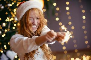 Encantadora chica pelirroja en un gorro de Papá Noel con bengalas en sus manos sonriendo a la cámara foto