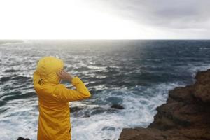 Mujer joven vestida con un impermeable amarillo de pie en el acantilado mirando las grandes olas del mar mientras disfruta del hermoso paisaje del mar en un día lluvioso en la playa de rocas en un clima primaveral nublado