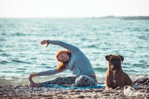 young woman doing yoga and stretching on the beach with her dog photo