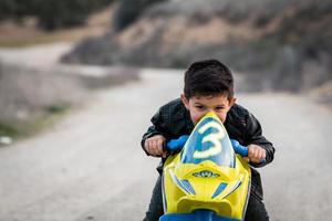 Un niño feliz conduciendo una motocicleta de juguete, vestido con una chaqueta de motociclista de cuero en una carretera rural foto