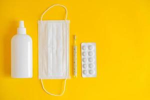 Un conjunto de mascarilla médica desechable, antiséptico, pastillas y un termómetro de mercurio sobre un fondo amarillo.
