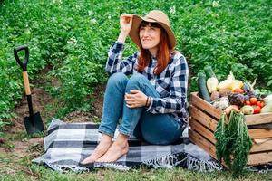 Mujer agricultora con una pala sentada sobre una manta cerca de verduras orgánicas frescas en una caja de madera con el telón de fondo de un huerto foto