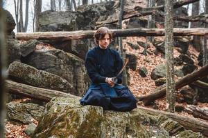 El hombre en kimono negro con una espada medita y se concentra en las rocas y el fondo del bosque foto