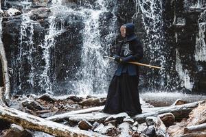 Hombre practicando kendo con espada de bambú en cascada, rocas y fondo de bosque foto