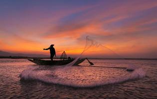 Asian fisherman on wooden boat casting a net for catching freshwater fish in nature river in the early morning before sunrise photo