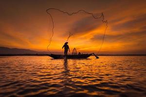 Pescador asiático en barco de madera echando una red para pescar peces de agua dulce en el río natural temprano en la mañana antes del amanecer foto