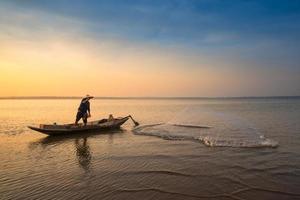 Asian fisherman on wooden boat throwing a net for catching freshwater fish in nature river in the early morning before sunrise photo