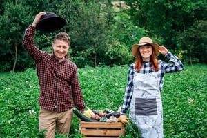 Man and woman farmers in hats holding fresh organic vegetables in a wooden box on background of a vegetable garden photo