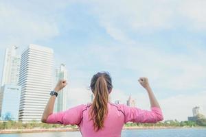 Back shot of young asian woman relax herself and warm down after city running exercise with a city view background and warm light cleared sky late of the afternoon. Outdoor running exercise concept. photo