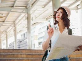 Happy Asian woman traveler tourist wanderer with trendy look searching direction on location map while traveling abroad in summer. Young female traveler with backpack and with map on street in city. photo