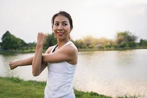 Mujer joven sana asiática del corredor que calienta el cuerpo que se estira antes del ejercicio y del yoga cerca del lago en el parque bajo la luz cálida de la mañana. estilo de vida fitness y mujeres activas se ejercitan en el concepto de ciudad urbana.