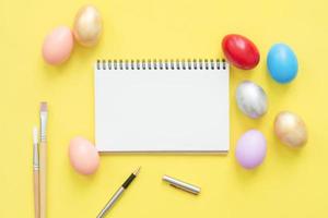 Flat lay top view colorful easter egg painted in pastel colors composition and mock up blank notebook with paint brush on yellow pastel color background. Easter day background top view with copy space photo