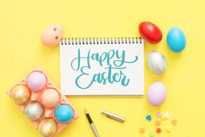 Flat lay top view colorful easter egg painted in pastel colors composition and text Happy Easter Day on notebook with paint brush on yellow pastel color background. Top view Easter day background. photo