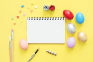 Flat lay top view colorful easter egg painted in pastel colors composition and mock up blank notebook with paint brush on yellow pastel color background. Easter day background top view with copy space photo