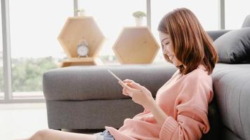 Asian woman using tablet while lying on home sofa in her living room. Happy female use tablet for texting, reading, watching video and surfing online at home. Lifestyle woman at home concept.