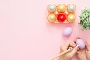 Top view person painting colorful easter egg painted in pastel colors composition with paint brush on pink pastel color background. Easter day background top view with copy space. photo