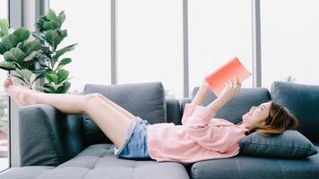 libro de lectura de la mujer asiática joven de vacaciones. un primer plano de la joven feliz acostado libro abierto en el sofá de casa. estilo de vida informal en casa. foto