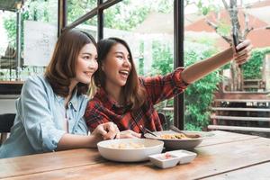 Hermosas mujeres asiáticas felices lesbianas pareja lgbt sentada a cada lado comiendo un plato de espaguetis con mariscos italianos y papas fritas en el restaurante o cafetería. pareja de mujeres con smartphone para selfie.