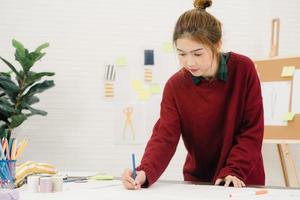 Hermosa diseñadora de moda asiática profesional que trabaja con bocetos de tela y diseño de ropa de dibujo en el estudio. concepto de trabajo de las mujeres de estilo de vida. foto