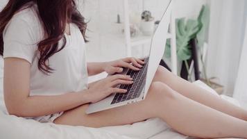 Beautiful Asian woman playing computer or laptop while lying on the bed in her bedroom. Happy female buying online shopping at home. Lifestyle woman at home concept. photo