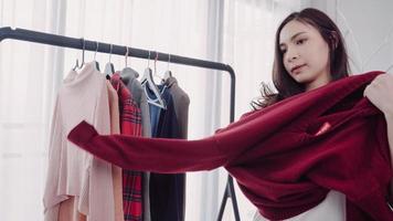 Home wardrobe or clothing shop changing room. Asian young woman choosing her fashion outfit clothes in closet at home or store. Girl think what to wear sweater.