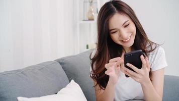 Asian woman playing smartphone while lying on home sofa in her living room. Happy female use phone for texting, reading, messaging and buying online at home. Lifestyle woman at home concept.