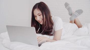 Portrait of beautiful attractive Asian woman using computer or laptop and listening music while lying on the bed when relax in her bedroom at home. Lifestyle women using relax time at home concept. photo
