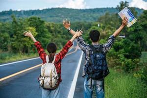 Both male and female tourists carry a backpack, stand on the road, and raise their hands on both sides. photo