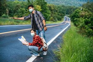 Female tourists sit and look at the map, male tourists pretending to hitchhike. Both wear masks and are on the side of the road. photo