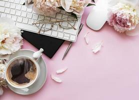 Flat lay top view women's office desk with flowers. Female workspace with laptop, flowers peonies, accessories, notebook, glasses, cup of coffee on pink background. photo