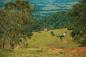 Pardinho, Brazil - May 31, 2018. Woman descending by cables in a sport called zip-line over meadows and trees in a valley near Pardinho. A small rural village in the countryside of Sao Paulo State. photo