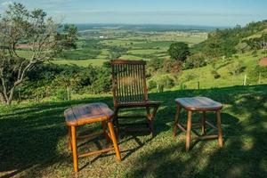 taburete y silla de madera a la sombra frente a un valle verde en un paisaje montañoso cubierto por bosques y praderas cerca de pardinho. un pequeño pueblo rural en el campo del estado de sao paulo. foto