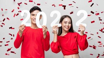 Lovely Asian couple smiling and showing number 2022 for new year concept on light gray studio background with confetti photo
