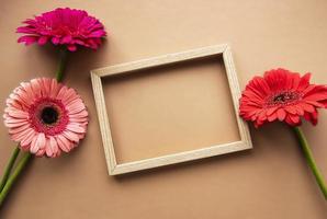 Wooden frame surrounded by beautiful gerbera flowers on a light brown background, top view, copy space, flat lay. photo