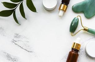 Rodillo de jade de masaje facial con producto cosmético sobre fondo de mármol blanco