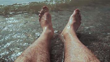 homem caucasiano sentado na água e as pernas lavadas pelas ondas do mar na praia video