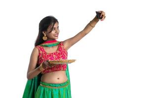 Beautiful young happy girl taking a selfie with clay lamp or diya during the festival of light Diwali using a smartphone on a white background photo