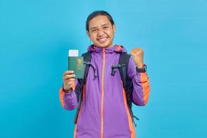 Portrait of cheerful asian young man showing parport book and making successful gesture isolated on blue background photo