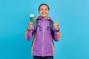 Portrait of cheerful asian young man showing parport book and making successful gesture isolated on blue background photo