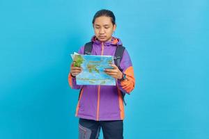retrato, de, asiático, joven, llevando, chaqueta, y, bolsa de transporte, mirar, serio, lectura, mapa, aislado, en, fondo azul foto
