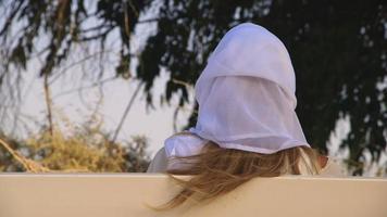 femme blonde vêtue d'une robe blanche et d'une coiffe blanche assise sur un banc dans une zone rurale. la femme assise dos à la caméra. ses cheveux flottent au vent. atmosphère champêtre de rêve video