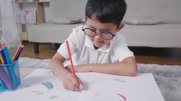 Vista frontal del niño feliz con gafas asiáticas pintando en un libro para colorear mientras está sentado en el piso en la sala de estar. feliz niño tailandés dibujando en papel en vacaciones en casa. concepto de relajación y afición. video