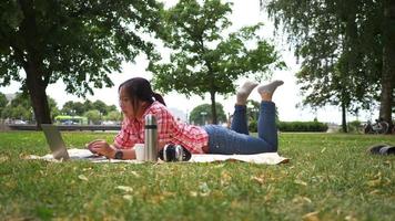 Aisan woman lying on mat and using laptop at the park. Opening and typing on laptop, working with good view at the park. Going out on the weekend. Going picnic concept