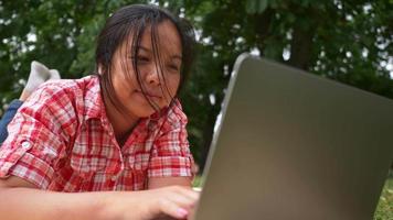 primo piano aisan donna sdraiata sul tappetino e utilizzando il computer portatile al parco. aprendo e digitando sul laptop, lavorando con una buona visuale del parco. uscire nel fine settimana. andando concetto di picnic video