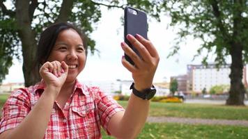 Close up Aisan woman sitting on mat and taking video call on smartphone at the park. Taking a rest with good view. Going out on the weekend. Going picnic concept