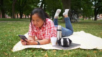 Asiatische Frau liegt auf der Matte und benutzt Smartphone im Park. bei guter Natur ausruhen, frische Luft schnappen. am Wochenende ausgehen. Picknick-Konzept gehen video