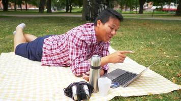 gelukkige aisan man liggend op de mat en met behulp van laptop in het park. videogesprek voeren op laptop met goed zicht op het park. uitgaan in het weekend. gaan picknicken concept video