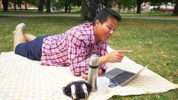 feliz aisan homem deitado na esteira e usando o laptop no parque. fazer videochamada no laptop com boa vista para o parque. saindo no fim de semana. conceito de piquenique indo video