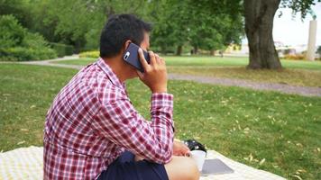 Happy Aisan man sitting, having a call on smartphone and holding a glass of water at the park. Taking a rest with good nature, getting some fresh air. Going out on the weekend. Going picnic concept video