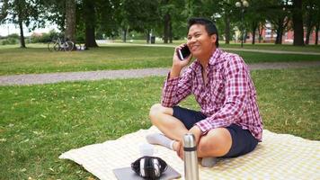 hombre feliz de aisan sentado en la alfombra, tener una llamada en el teléfono inteligente y beber agua en el parque. tomar un descanso con buena naturaleza, tomar un poco de aire fresco. salir el fin de semana. ir a concepto de picnic video